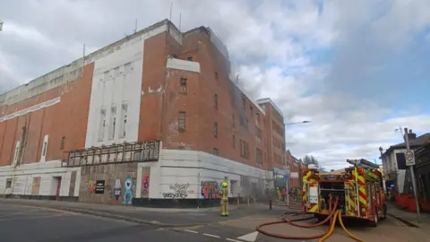 London Fire Brigade A fire engine with hoses connected to the back stands at the side of a tall brick building with smoke coming out of the upper windows. 