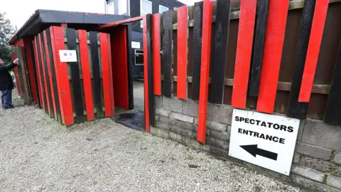 Getty Images A fence with red and black panels. A sign indicating the way to a 'spectators entrance' sits on the wall beneath it.