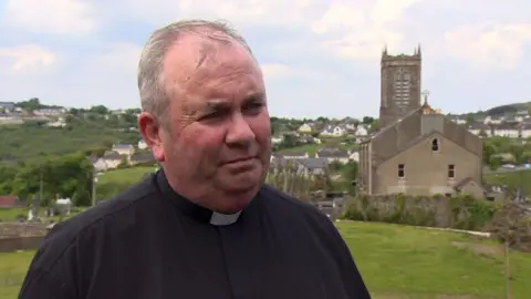 A priest, with short grey hair, looking at the camera. There is a field and a church in the background.