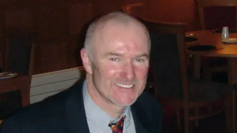 Dr Mohammad Husban Colin Howell is smiling at the camera. His hair is closely shaved and there is some grey stubble on his chin. He has a ruddy complexion and is wearing a suit with a bright tie.