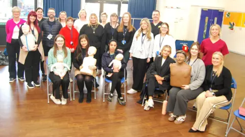 KCH A large group of people in a room, with six sitting down and five people holding dummies. They are in a room with a wooden floor, blue curtains, and windows. Some are sat down on chairs and the rest are standing up. 