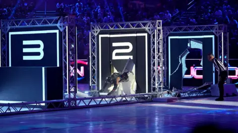 BBC/Hungry Bear Action shot of the show Gladiators, featuring a contender breaking through a wall.