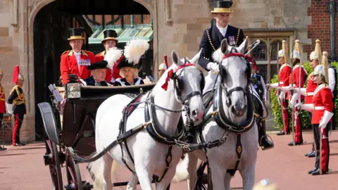Reuters King Charles and Queen Camilla ride in a carriage after the Order of the Garter service. Two footmen sit behind them. White horses pull the carriage with a single rider. They are flanked by a row of guards.