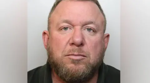 Avon & Somerset Police A police custody head and shoulders mugshot of Mark Killick. He has a scruffy dark brown beard and receding hair on his head and is wearing a dark coloured polo top
