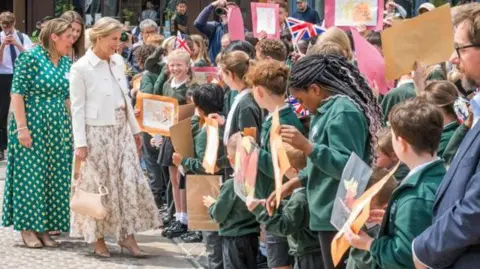 West Northamptonshire Council A blonde haired woman in a cream coloured outfit pictured talking to a row of students in green blazers holding up an eclectic mix of art work. Union Jack flags can be seen in the background