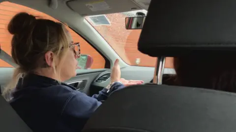 Seb Cheer/BBC A woman sits in the passenger seat of a car as it drives along. She is acting out simple signs with her hands, for the driver to observe.