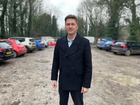 Man in black coat and brown shoes stands in a gravel car park surrounded by parked cars and trees 