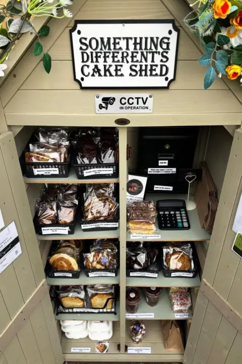 A beige coloured cake shed. The doors are open and there are 10 shelves stocked with cakes. A calculator is on the top shelf and there is a sign which reads Something Different's Cake Shed at the top, along with a CCTV sticker.