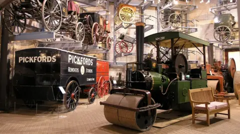 Stockwood Discovery Centre A collection of horse-drawn carriages with some on raised platforms by the wall in a museum. There are also two steam rollers in the middle.
