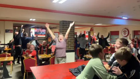 Fans inside a building are stood up and cheering with their arms in the air. 