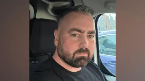 Family photograph A composite image of a man sat in the seat of a car. He has slicked back grey hair and a well-trimmed dark beard and is wearing a black t-shirt.
