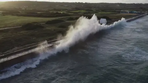 BBC A large wave crashing up causing a large splash of foam during high tide at St Ouen. An expanse of green fields can be seen.