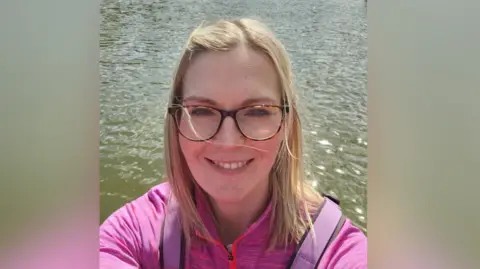 Linda Rukin The picture shows the head and shoulders of Linda Rukin who has shoulder length blonde hair. She is wearing glasses and has a pink top on and behind her is a lake. 