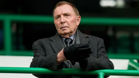 Sir Tom Farmer standing in a football stand. he is wearing a greay coat and grey shirt and tie