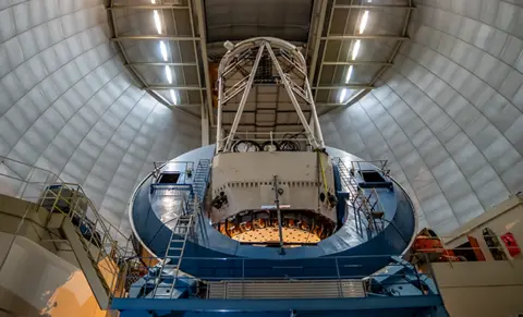 DESI A blue telescope inside a dome. It consists of a square base on which sits a circular ring from which a thick cream coloured antennae protrudes.