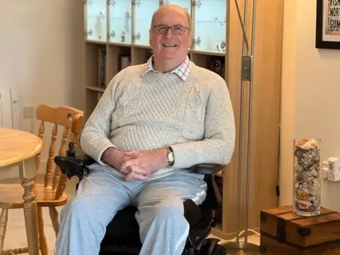 Martin Wallace wearing a cream jumper and white trousers, sitting in a wheelchair in his living room.