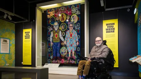 A man sits in a wheelchair next to a tapestry in a museum. The tapestry depicts an elderly couple holding hands. A yellow information board headlined, 'Wish you were here' is behind him.