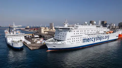 Mercy Ships Two large white hospital ships are docked side by side in a busy port. One is the Global Mercy and the other is the Africa Mercy, with tents and port buildings nearby.