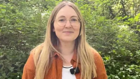 Becca is standing in front of a green hedge in the sun. She is wearing an orange cardigan and has long, blonde hair and glasses.