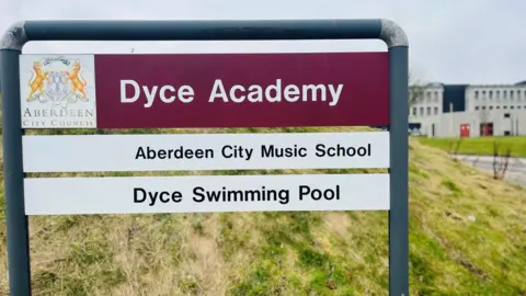 Dyce Academy school sign, with school building in the background.