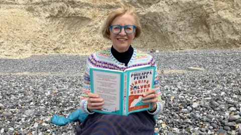 A woman sitting on a pebbled beach in front of cliffs with an open book in her hands. She is wearing a colourful knitted cardigan and blue framed glasses, and looking at the camera.