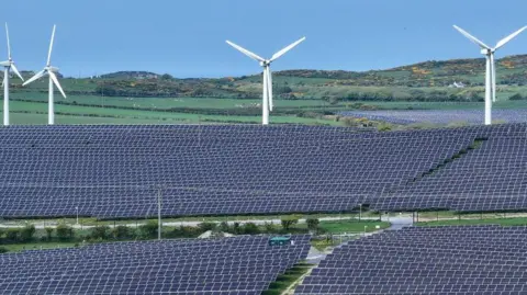 Getty Images Several fields obscured by hundreds of solar panels with 4 wind turbines in the background and rolling hills further in the distance. 