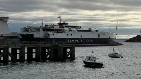 BBC The Manannan, a sleek catamaran which is red, white and black and has Steam-Packet.com on the side in white lettering, as it leaves Douglas Harbour.