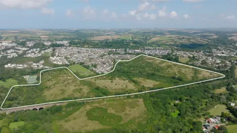 An aerial view of the site in Dudman Farm site in Cornwall. It is a large plot of land, including fields and trees, with some boundary housing 