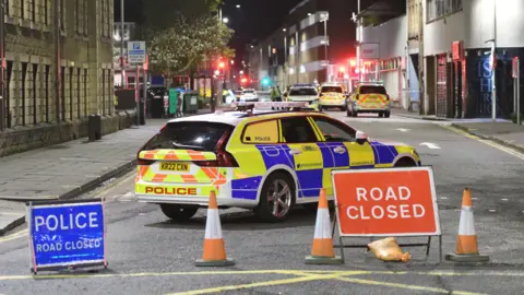 Stuart Cowper A police car sits behind two road closed signs and three traffic cones. Other police vehicles are seen in the distance