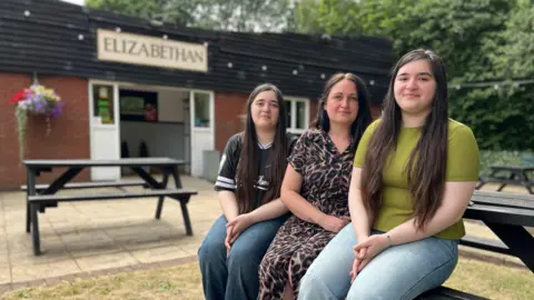 Landlady Kely Lake sits on a picnic bench outside the pub in between her twin girls, with Peyton on the left and Tamsin on the right. The Elizabethan is a one storey building with red bricks and wooden cladding. A hanging basket and outdoor lights can be seen on the front of the pub. The twins both have long brown hair and are wearing short sleeved tops and jeans. Kely also has long brown hair and is wearing a leopard print dress.