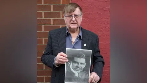 Robin Maudsley Robin Maudsley has brown combover hair with white sideburns and spectacles. He wears a dark blue pinstripe jacket blue shirt and holds photo of his cousin.