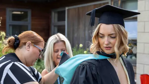 A woman with curled blonde shoulder length hair is looking towards the ground and wearing a light yellow dress and gold necklace underneath a black graduation gown and cap. The back of the gown is blue and two women are smiling as they sort it out for her. You can see a brown building and shrubbery in the background.