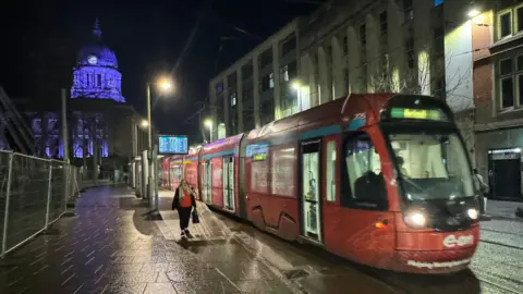 Night-time photo of a red tram travelling through Notttingham city centre with the Council House visible in the background