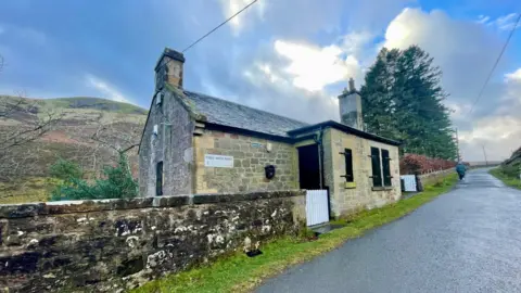 A paved road passes through the photo next to Loganlea Cottage. It is built of stone and has stone walls. There are some trees and a hill behind it.