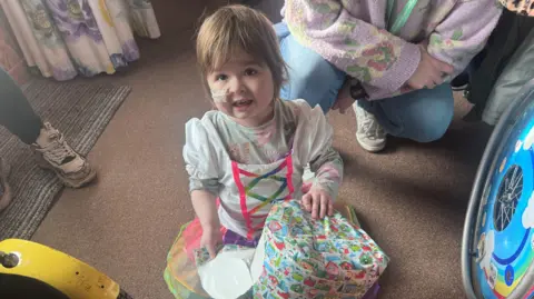 A young girl in a tutu dress opening a present. She has a tube going into her nose.