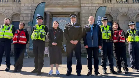 Nine people stand in a line, some police officers wearing yellow high vis vests, and some city officers wearing red vests.