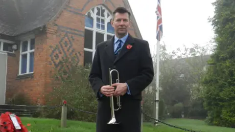 A man in a dark coat and suit and poppy, holding a bugle, standing beside a memorial with poppies on it. Behind him is a red brick building with some darker bricks in a diamond pattern. A big tree is to the right of the image with a flag pole with the union flag on it, next to it.