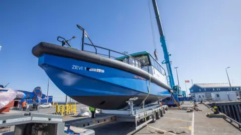 A bright blue electric boat called Zevi 1 sits on top of a wheeled metal trolley on dry land