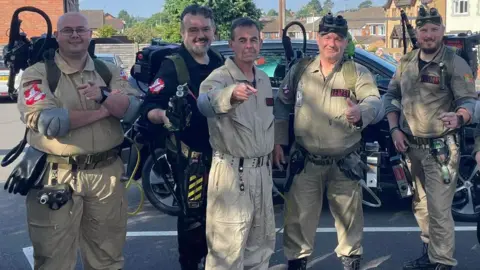 Steve Robbins There are five men lined up in a parking lot. They wear all-in-one Ghostbusters suits with proton packs on their backs and smile at the camera. 