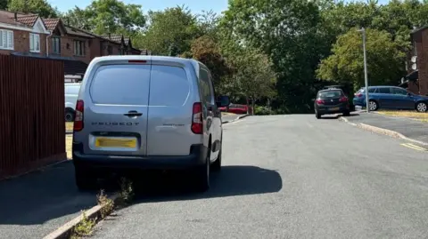 Charlie Stubbs / BBC There is a silver Peugeot van parked on the pavement, leaving a tight space for people to get around. The license plate is blurred. In the background, there is a black car that has also parked itself on the pavement.