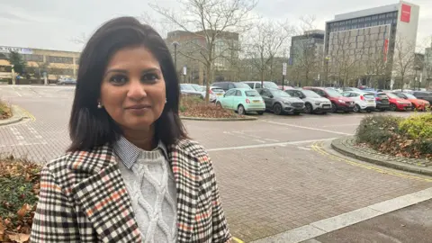 A picture of a woman with dark brown hair. She is wearing a white and black striped jacket with an orange stripe too. She is also wearing a white jumper underneath and is stood ina car park with buildings behind her.