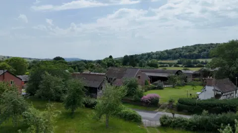 The farm includes historic timber barns, and modern buildings. There is a black and white cottage and an orchard next to the old barns.