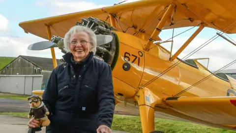 Betty Bromage stood in front of a yellow plane. She has short white hair and is wearing glasses and a dark coat. She is holding a teddy bear and smiling.