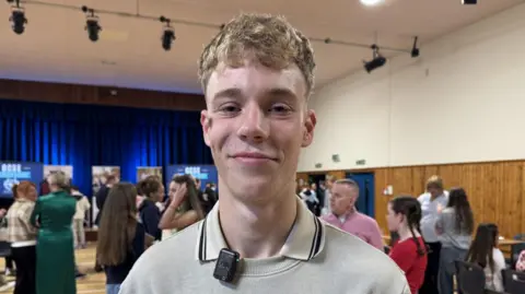 Max is wearing a dull neutral coloured jumper with a polo collar sticking over his round neck collar. A busy assembly hall is behind him.