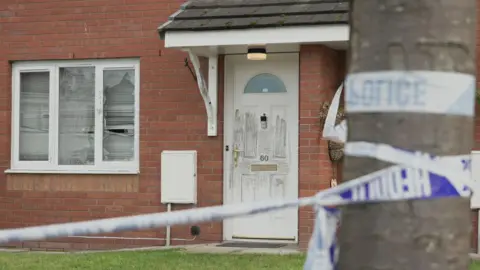 Police tape tied to a tree outside a red-brick house with a porch and a white door.