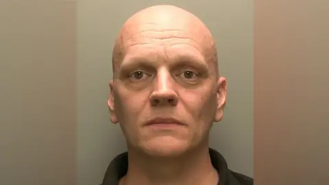 Ben Cairns has brown eyes and is clean shaven with no hair on his head.