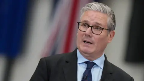 Sir Keir Starmer speaks, wearing spectacles, a dark suit, light blue shirt and a navy blue tie, against a blurred background