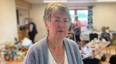 A woman with short grey hair, glasses, a white top and a pale blue cardigan in a room with tables and people blurred in the background. 