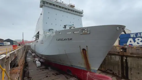 A large grey ship with the words “Cardigan Bay” painted on its side sits in a dry dock. A red metal gangplank connects the ship to shore in the distance.