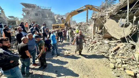 BBC/Goktay Koraltan A large group of people stand in front of a destroyed building. In the background a digger moves pieces of the building.